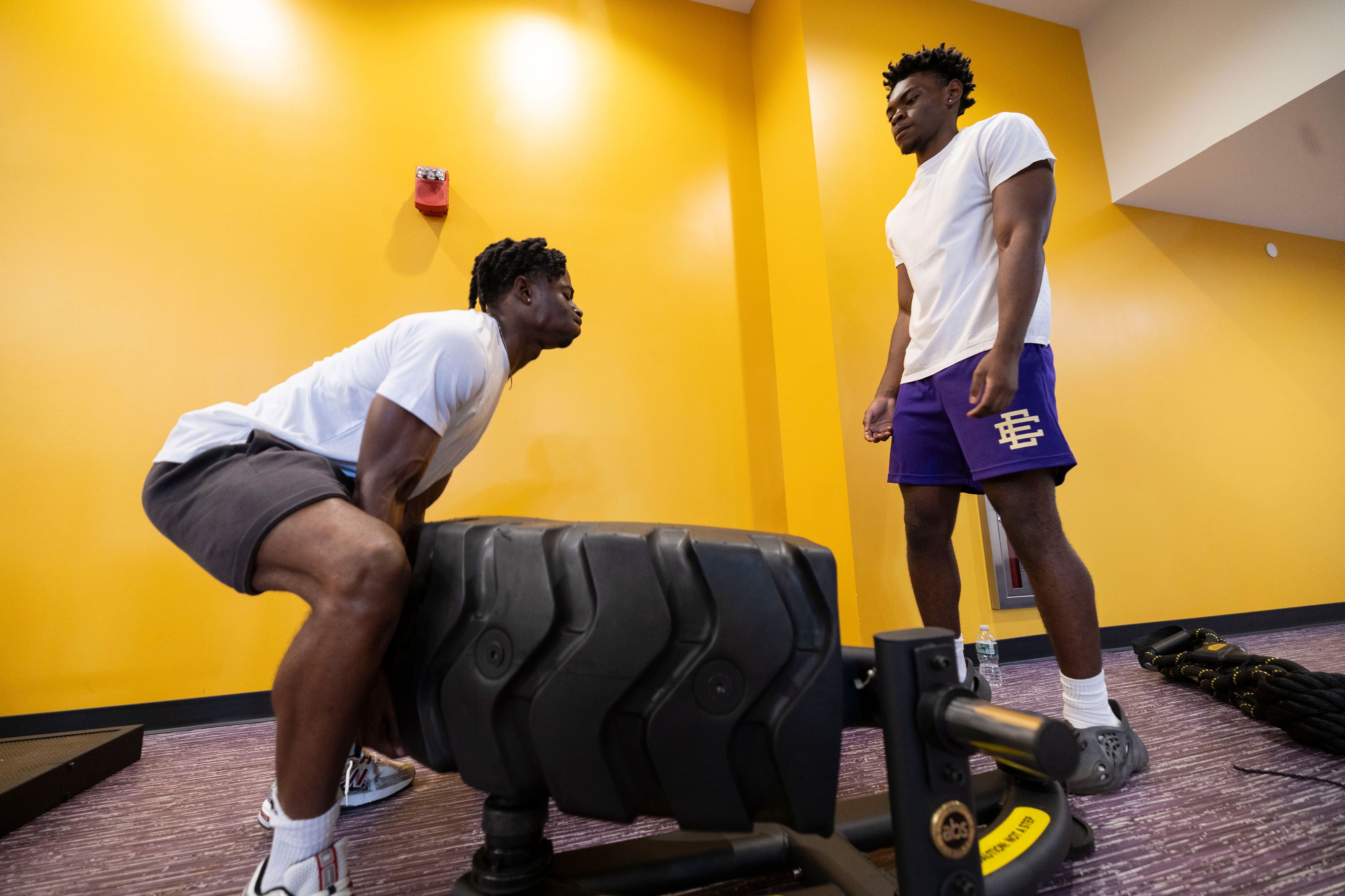 A student squats and lifts half a industrial tire inside a workout room while another student spots him.