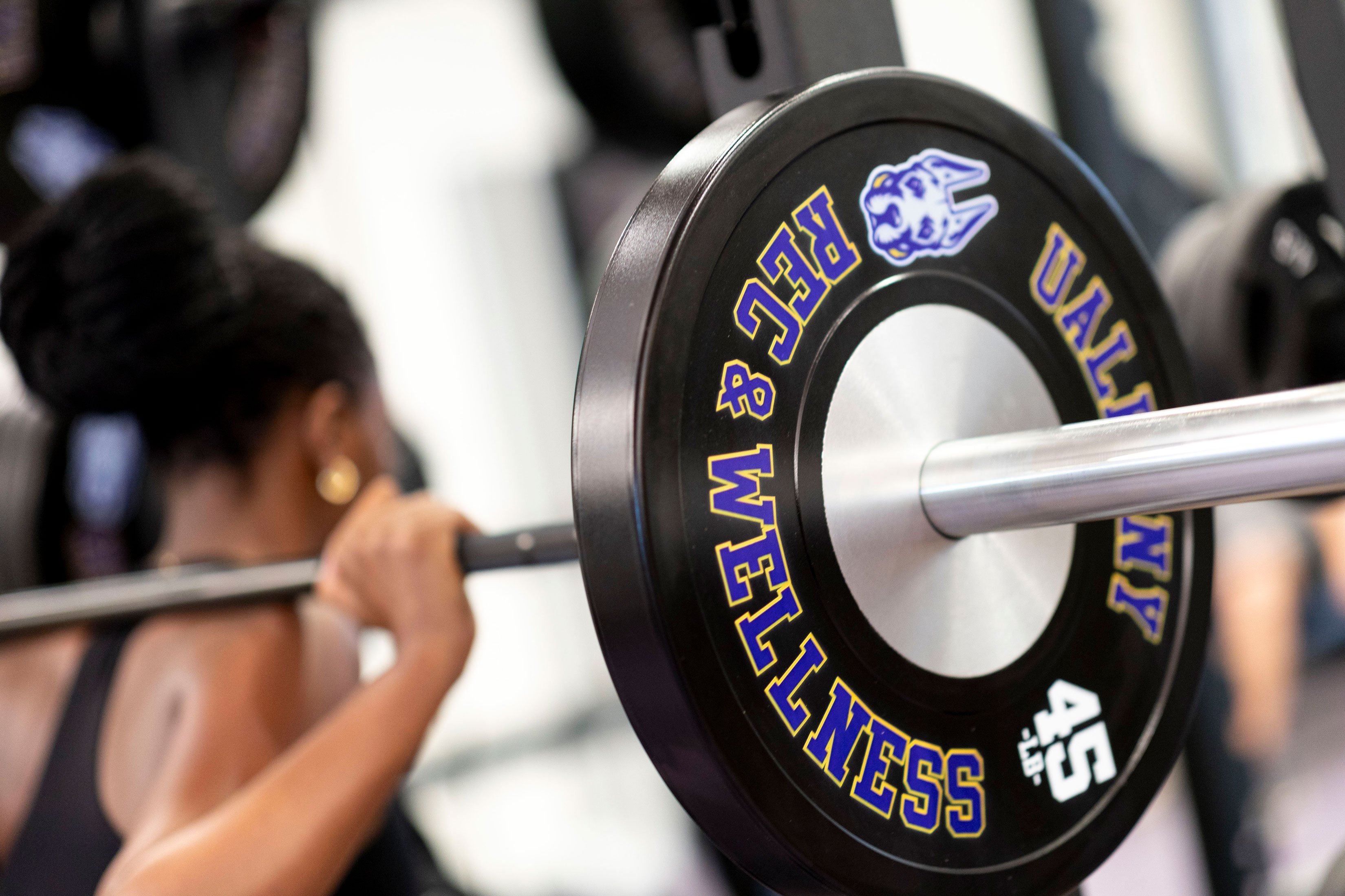 A student squats while holding a bar with weights on her shoulders. The weights say "Rec & Wellness" and have an icon depicting UAlbany's mascot Damien the Great Dane.