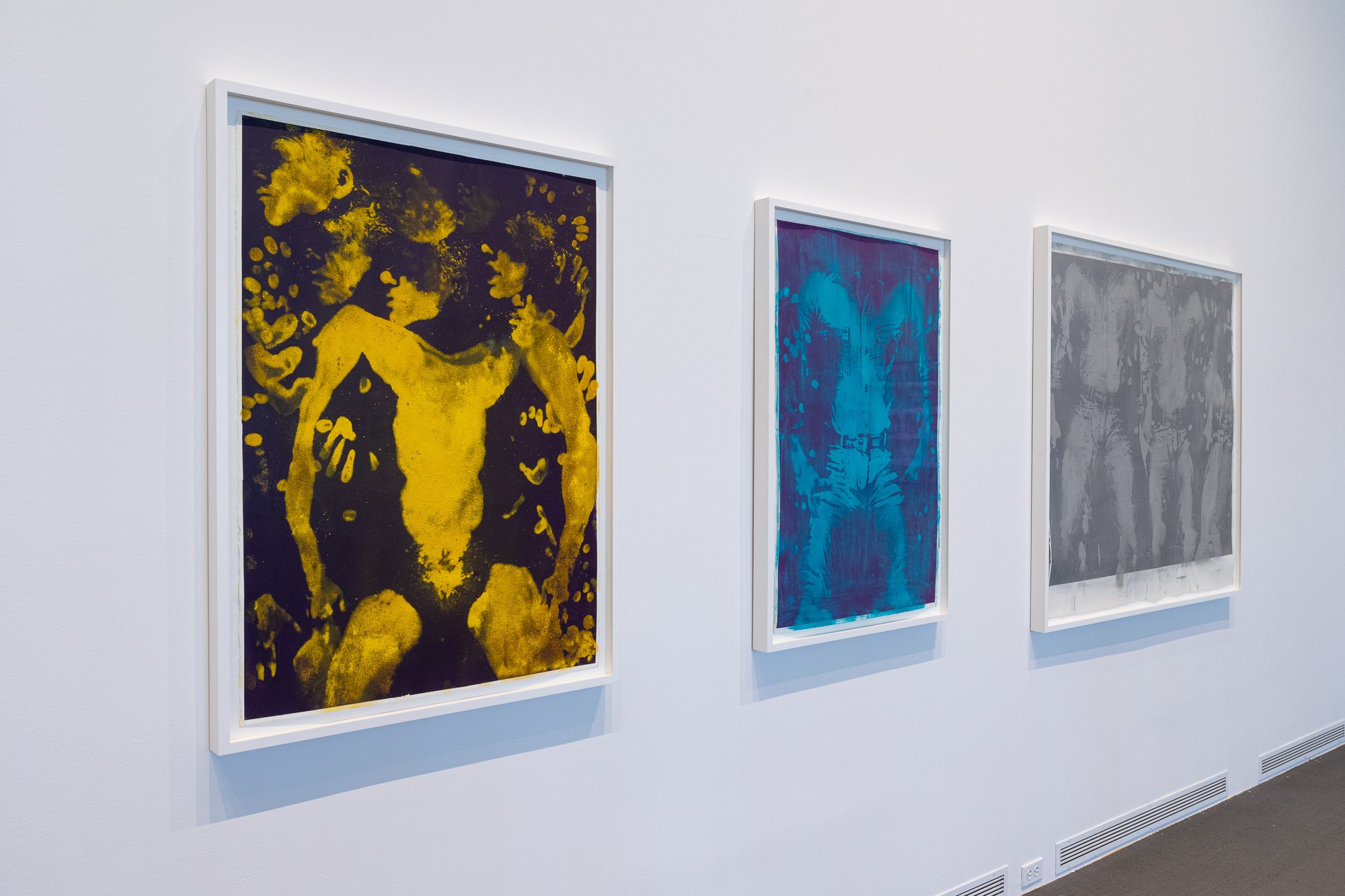 Angled image of three framed prints of bodies. The first is in yellow and black, the second features shades of blue, and the third includes shades of gray.