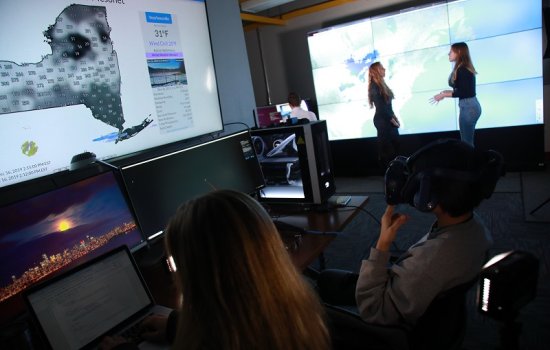 Two people sit a screens in a darkened computer lab, with one of them wearing VR goggles, as to people speak in the background.