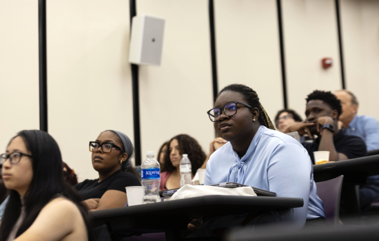 Students sit at desks in a lecture hall and look toward the front of the room.