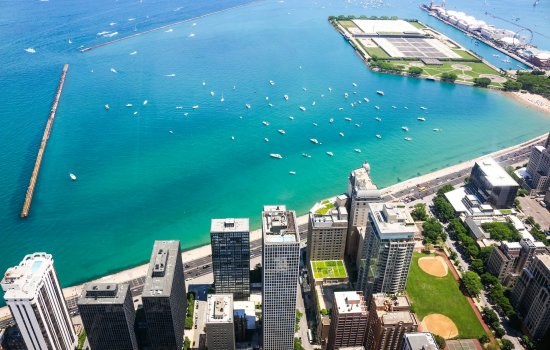 Aerial image of the Chicago shoreline along Lake Michigan. The bottom third of the image shows city buildings with tree lined streets and two baseball diamonds. The top two-thirds of the image is aqua blue water, dotted with boats, two jetties and two fingers of land reaching out into the lake. One has an amusement park and Ferris wheel. The other has a mix of parkland and water purification infrastructure. 