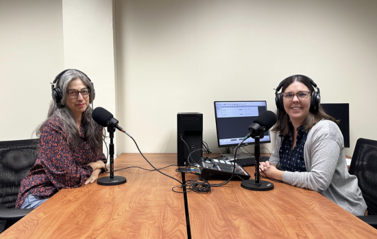 Two women wearing headphones sit in front of microphones at a table next to a computer and recording equipment.