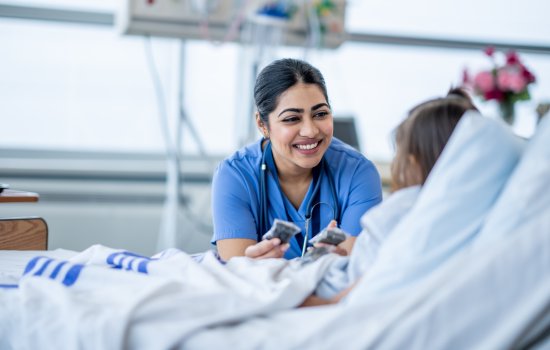 A female nurse wearing blue scrubs and a stethoscope smiles as she interacts with a pediatric patient reclined in a white hospital bed.