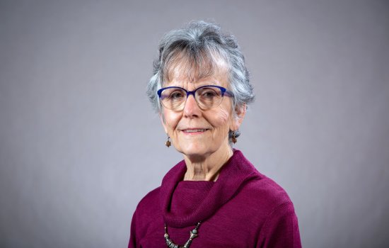 A woman with short gray hair and glasses wears a pink sweater and poses for a portrait against a gray backdrop