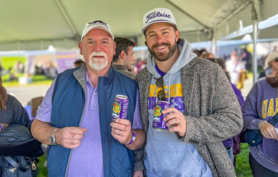 Two men with beards and white ball caps stand in a tend holding purple cans of Reed's ginger beer, decorated with a white mustache.