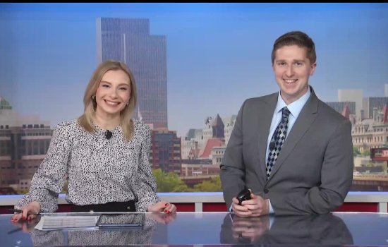 UAlbany alum Reid Kisselback and a female colleague smile and pose while sitting behind a newscasters desk. The Albany skyline is projected behind them.
