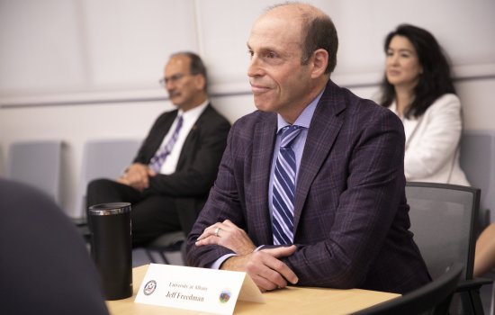 A man in a suit and tie sits at a table with a name card that reads "University at Albany Jeff Freedman."