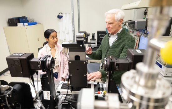 A young female student and an older male professor talk amidst numerous high-tech devices in a lab room with a filing cabinet in the background and devices in the foreground.