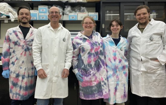 Five people, two women and three men, stand together smiling for a group portrait in front of a lab bench. All are wearing lab coats, three of which are tie dyed blue, pink and purple. Shelves of lab supplies can be seen behind them.