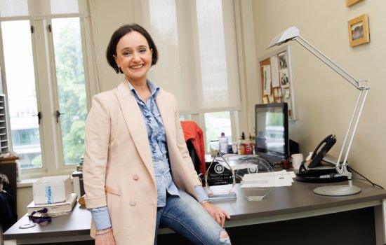 A woman with short brown hair wearing a blue blouse, cream blazer and jeans is smiling at the camera while seated on a desk.