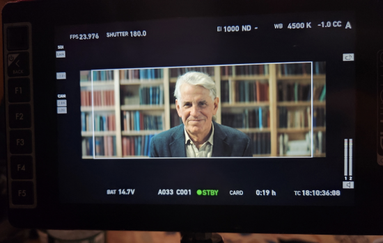 A man with short white hair in a collared shirt and blazer appears on a film screen seated in front of wall-to-wall bookcases.