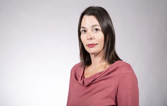 A woman with shoulder-length brown hair in a pink top poses for a portrait against a gray backdrop.