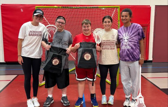 A group of three women wearing Lacrossing Barriers shirts stand with two men with special needs in front of a lacrosse net inside of a gym. A woman at the far right of the photo wears a UAlbany lacross jersey.