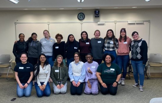 A group of 16 members of STEM NOW pose, all smiling, for a group photo in a classroom at UAlbany. The group is posed in front of a white board and they are arranged in two rows. The front row is kneeling on the carpet, which is gray. All are wearing white name tags.