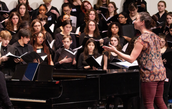 Young people dressed in all black are holding music books and singing on a stage with a piano and conductor.