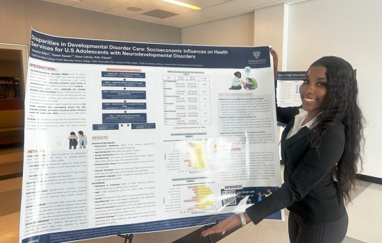 A young woman with long dark hair, wearing a black sweater and dress slacks, smiles as she displays her poster titled "Disparities in Developmental Disorder Care: Socioeconomic Influences on Health Services for U.S. Adolescents with Neurodevelopmental Disorders."