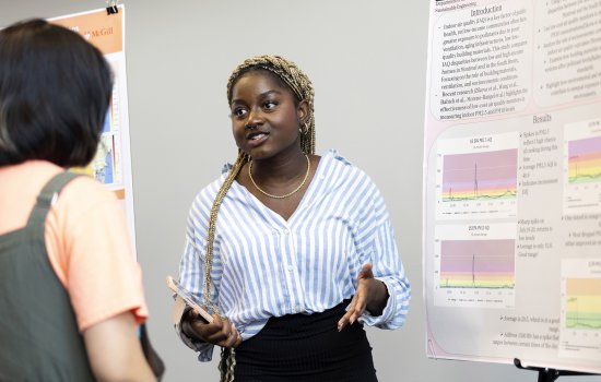 An African -American young woman with a white and blue vertical-striped blouse, a gold necklace and a black skirt gestures with her hands while speaking in front of an academic poster indoors.