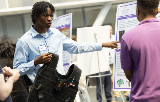 A man with short black braids in a light blue shirt holds up a black vest and gestures to a poster as he speaks to a man standing nearby in a purple shirt