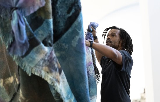 An African American man holds a large colorful tapestry from one end as it is lifted up at the other end in a room with white walls.