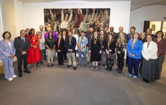 More than 2 dozen smiling people, many wearing medalions, stand in a semicircle in front of a large painting