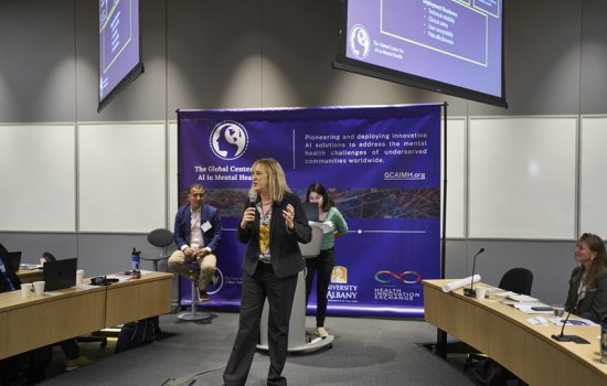 A woman in a black business suit stands holding a microphone in front of a purple banner.