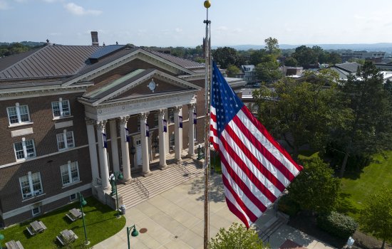 The American Flag is seen in the foreground on a flag pole in front of a colonial-style college building against a blue sky with green lawn and trees in the background.