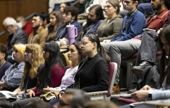 Rows of people sit in auditorium-style seats, listening intently to a speaker at the front of the room.