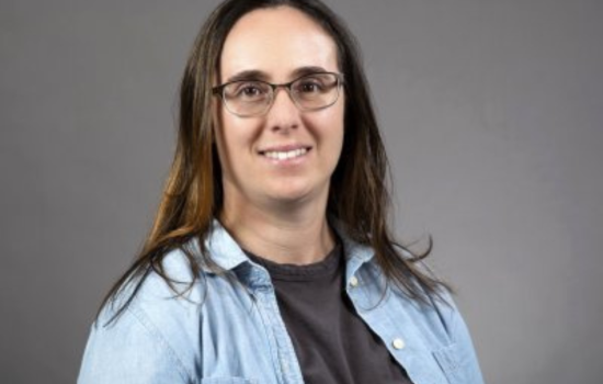 A woman wearing glasses and a denim shirt smiles at the camera