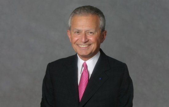 A man with silver hair in a black suit and pink tie smiles for a portrait against a gray backdrop.
