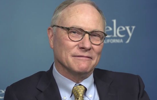 A man with balding hair and glasses, wearing a dark suit and yellow/gold checkered tie sits for a portrait with a logo of the University of California, Berkeley behind him.