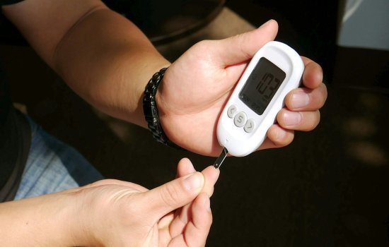 A person, seated, holds a blood sugar monitor, pressing the metal test tip to the tip of their right index finger.