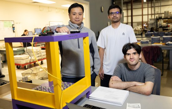 Three young men appear with a gold and purple device with lots of buttons on it. One of the men is seated folding his arms. The device has a large button on top.
