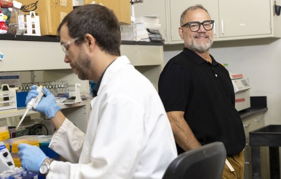 In the foreground, a man wearing a white lab coat and clear protective glasses handles a pipette. To the right, standing behind him is a man wearing a black polo shirt and black square rimmed glasses.