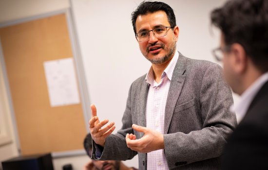 A man with dark hair and a gray blazer and glasses speaks animatedly in a brightly lit classroom.