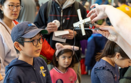 A young boy in glasses and a babseball cap and a young girl in a headband watch as someone out of frame spins a windmill made from Legos.