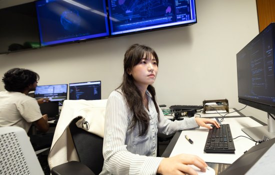A female student with dark long hair and brown eyes wearing a striped blouse sits at a computer terminal with screens in the background displaying a digital globe in blue type.
