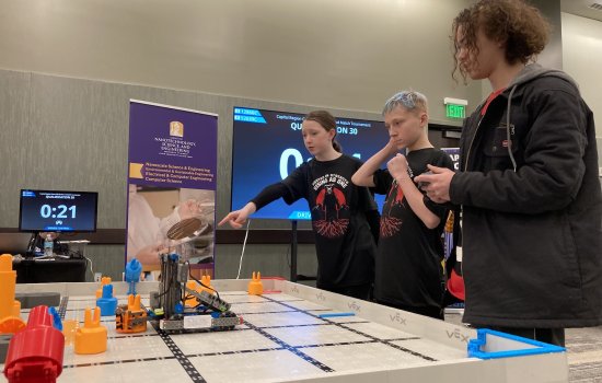 Girl points as two boys stare down at a robotic forklift on a game board.