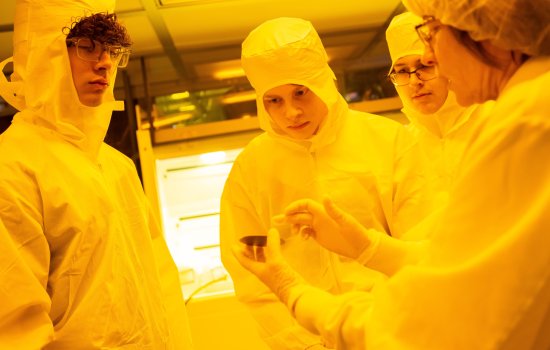Three students in protective bunny suits examine a semiconductor wafer held by an expert.
