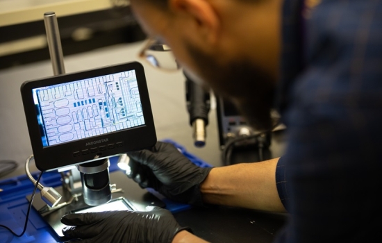 A blurred man in the foreground wearing glasses and black gloves leans toward a black screen displaying electronically magnified computer circuitry.