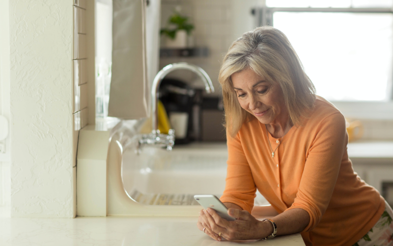 A woman wearing an orange shirt looks at her cell phone in her kitchen. 