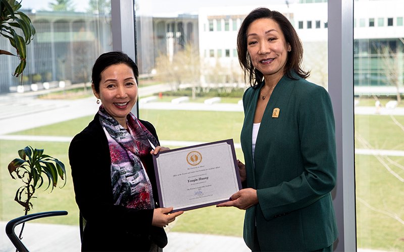 Youqin Huang stands left in a black jacket and patterned scarf while accepting a certificate from UAlbany Provost Carol Kim, right, wearing a green blazer with a gold pin. They are smiling in front of a window with a view of campus buildings and grass.