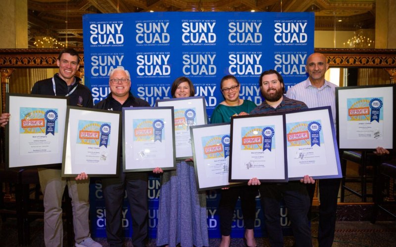 Six smiling people hold up eight large, framed awards certificates