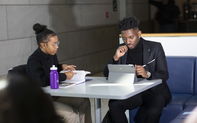 A young woman and man study together at a table, the woman with an open book and the an with a tablet computer