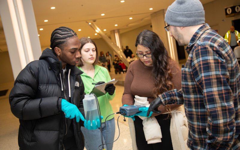 Four young people, two men and two women, stand together holding instruments to collect and record water quality measurements. One of the women is holding a gray electronic device connected to wires with probes placed in a water bottle held by a man wearing blue gloves. The second woman looks on, holding a clipboard and pen. The second man, wearing a plaid shirt instructs the group. 
