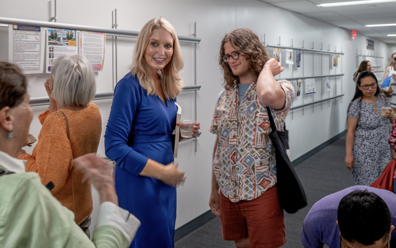 A woman with long blonde hair in a blue dress smiles in a hallway full of people who are viewing artwork on the walls.
