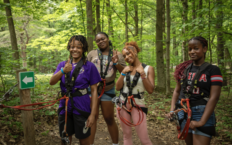 Four students wear harness gear for a high ropes course and pose for a picture in the woods.
