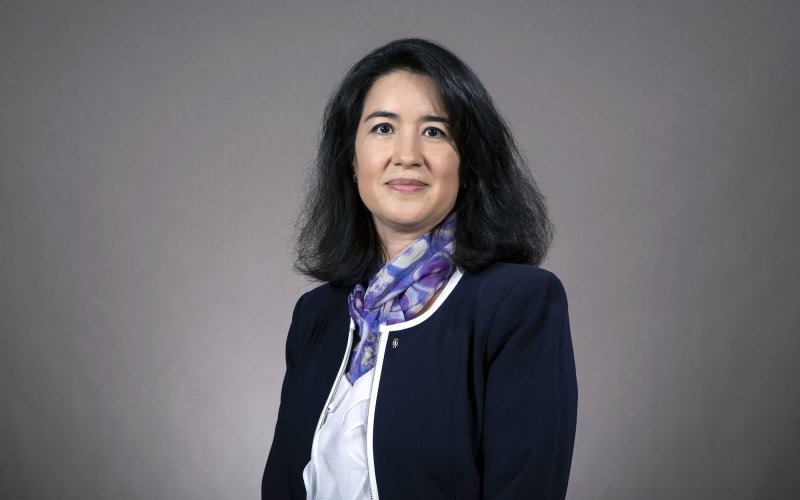 A woman with shoulder-length black hair wears a blue and violet scarf over a white blouse and navy blue blazer, and poses for a portrait against a gray backdrop.