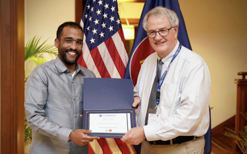 Two men smile and jointly hold a Fulbright certificate as they pose for a photograph in front of two flags.