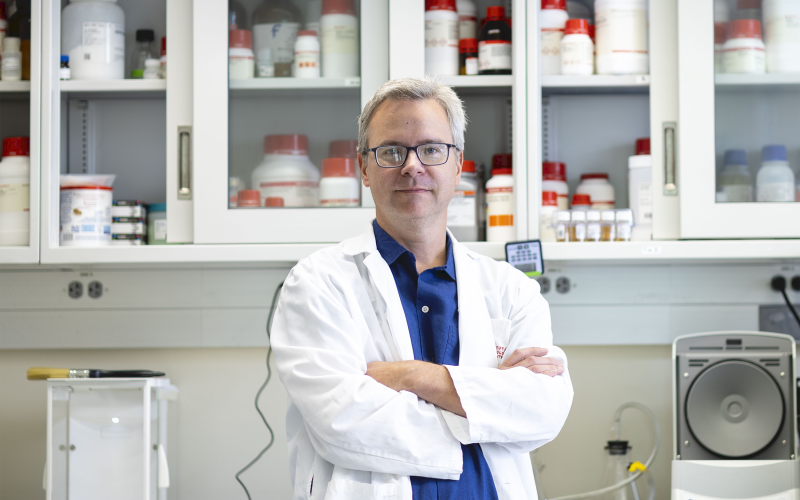 Associate Professor of Nanobioscience Ben Boivin wearing a white lab coat and blue shirt and glasses stands in his lab in front of a cabinet with clear glass panels and numerous white bottles with red caps.